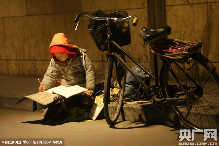 鄭州中原路京廣鐵路大橋下涵洞的馬路牙子邊，一位戴著紅色辣椒帽的小男孩趴在一個(gè)小馬扎上，上面鋪著一張車墊，在微弱的燈光下寫作業(yè)